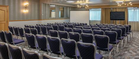 A conference room with rows of chairs, a projector screen, and a large flat-screen TV on the wall.