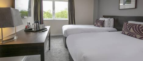 A hotel room with two beds, a desk with a lamp, a coffee maker, and a window with curtains.