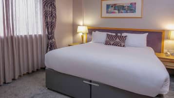 Habitación de hotel con una cama grande, una mesita de noche con lámpara, un cuadro enmarcado en la pared y una ventana con cortinas.