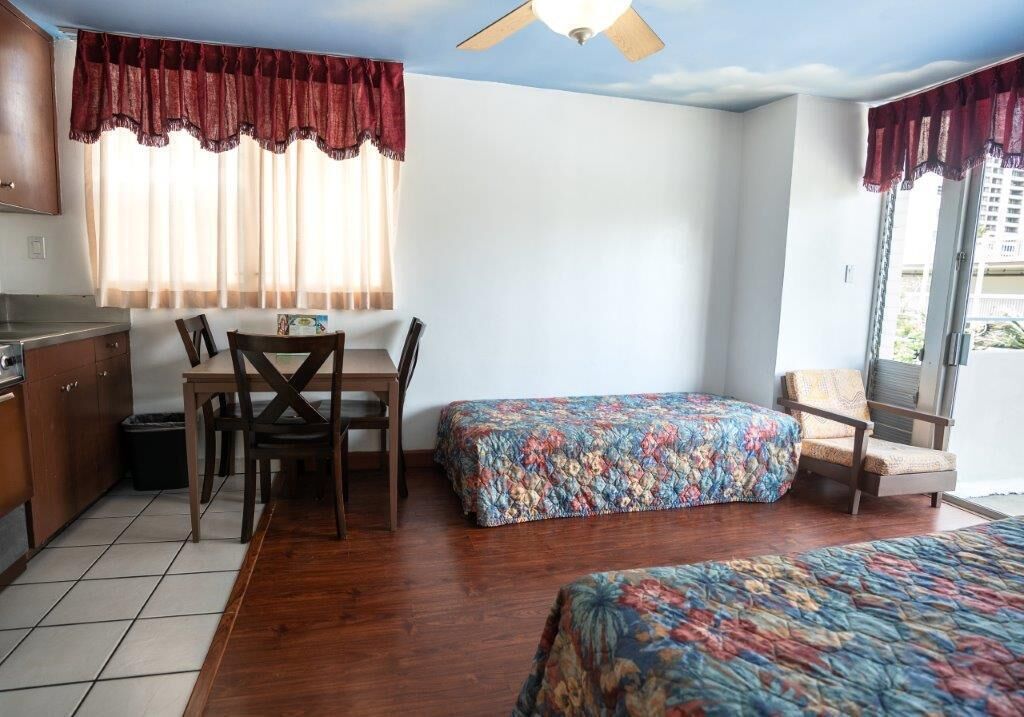 Quarto com mesa de jantar, duas cadeiras, cama com colcha florida, ventilador de teto e janela com cortinas.