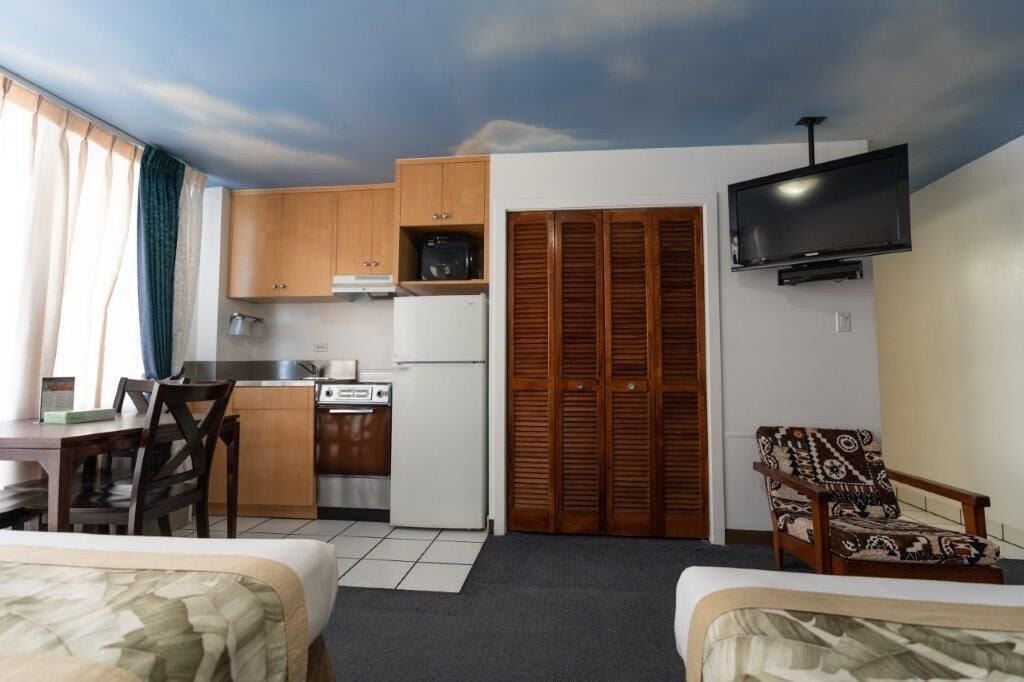 A compact hotel room with a kitchenette, a TV, a bed, and a chair.