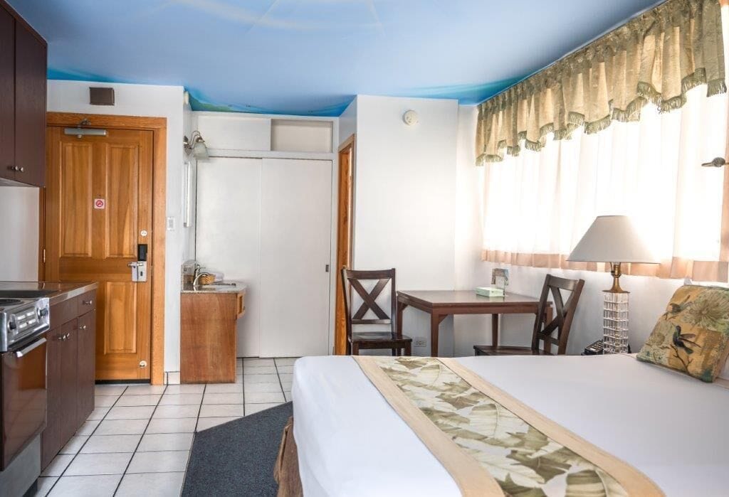 A hotel room with a bed, a desk, a chair, a small kitchenette, and a window with curtains.