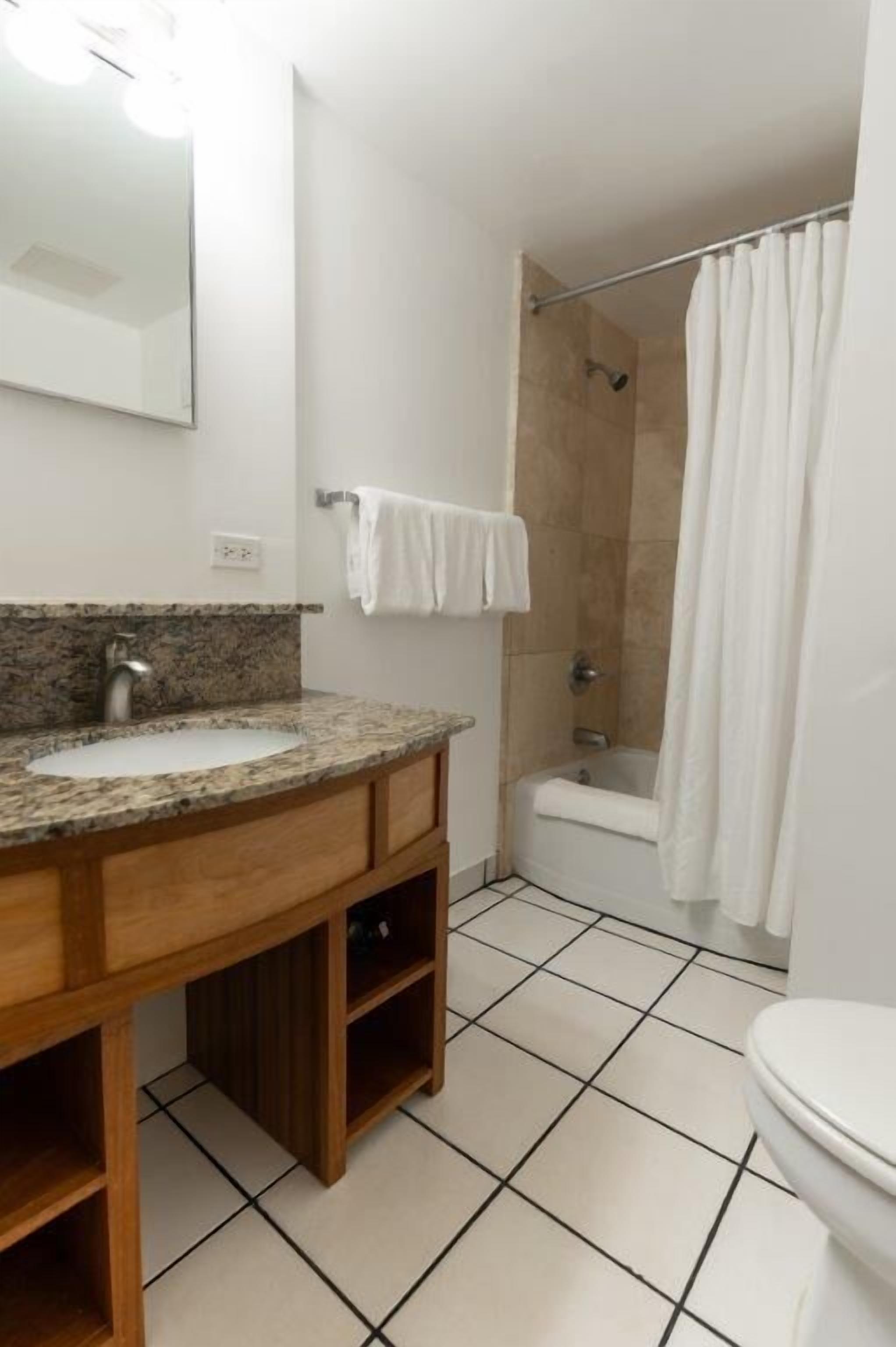 Een badkamer met een wastafel met granieten blad, een houten kastje, een wit toilet en een douche met een witte gordijn.