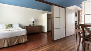 A bedroom with a wooden floor, a bed with a white bedspread, a nightstand with a lamp, and a sliding door closet. - Holiday Surf Hotel (Honolulu)
