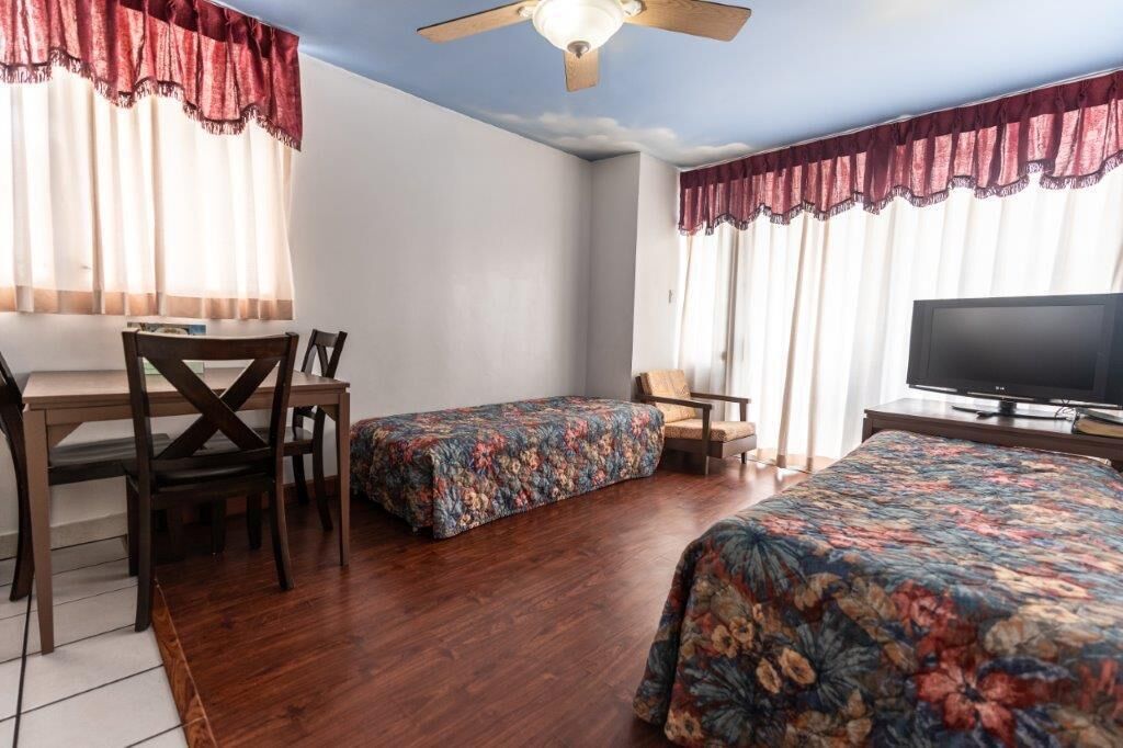 Una habitación con dos camas individuales, televisión, ventilador de techo, una mesa con sillas y cortinas.