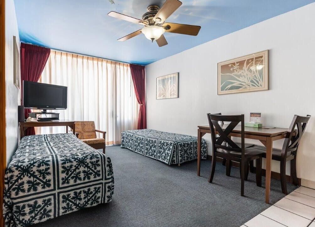 A hotel room with two beds, a TV, a ceiling fan, a dining table with chairs, and artwork on the walls.