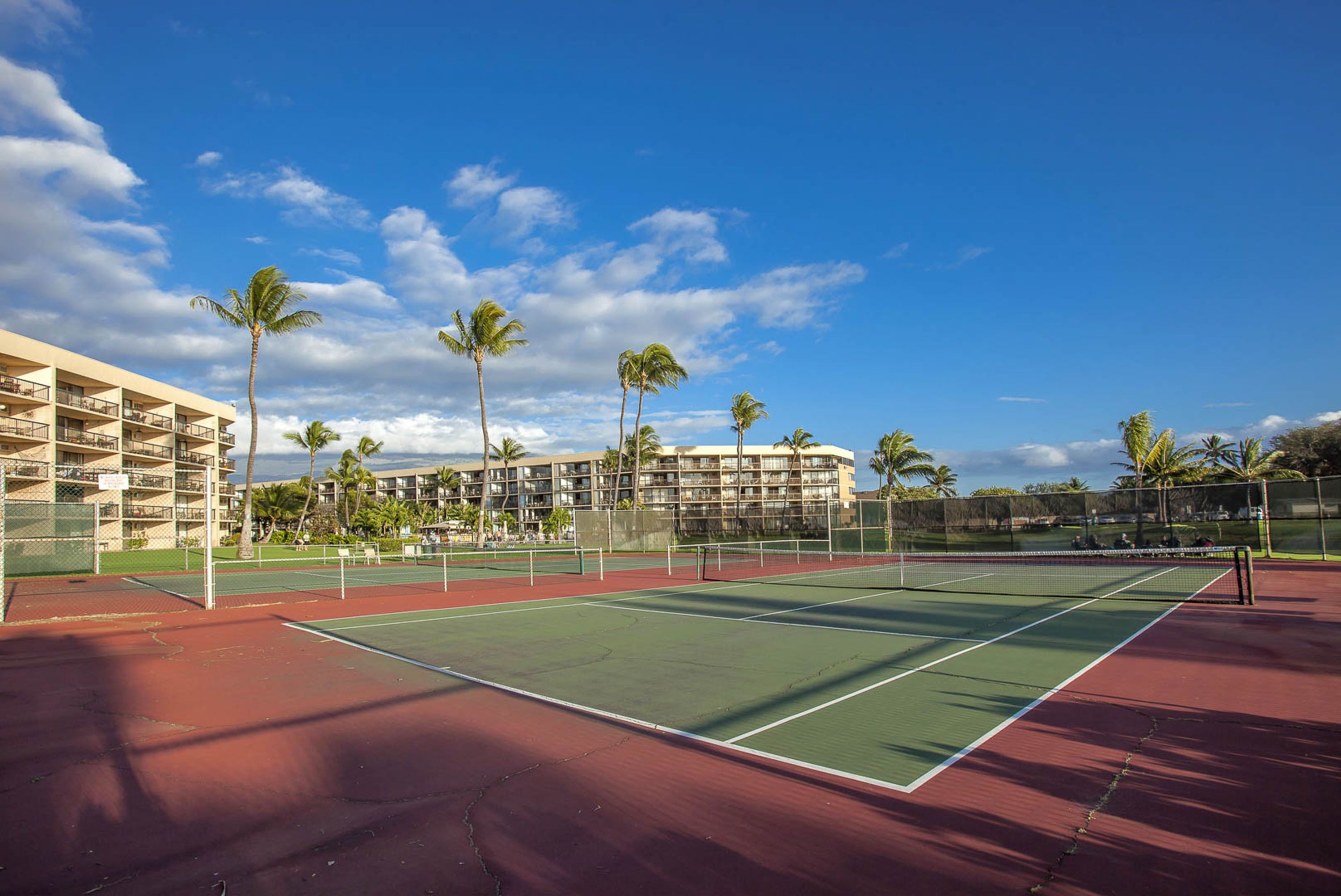 An outdoor tennis court with a red and green surface, surrounded by palm trees and a hotel building in the background.