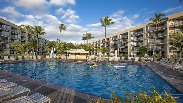 A hotel pool area with multiple lounge chairs, palm trees, and a clear blue sky. - Maui Sunset - Maui Condo & Home (Kihei)