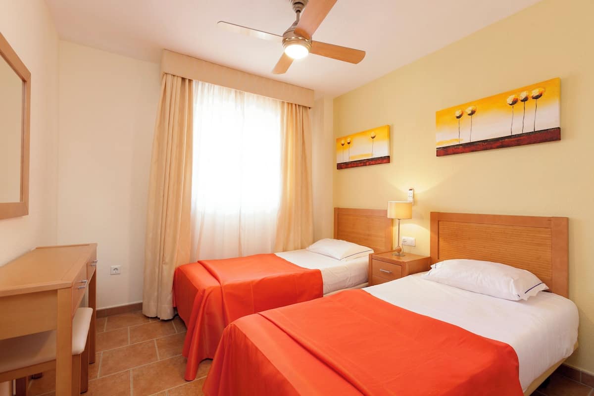 Habitación de hotel con dos camas, un escritorio, un ventilador de techo y una ventana con cortinas.