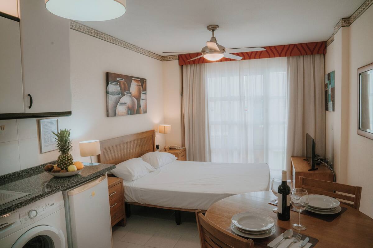 Habitación de hotel con cama, una mesa de comedor con platos y vasos, lavadora, ventilador de techo y un cuadro en la pared.