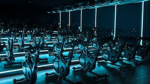 Un gimnasio con bicicletas estáticas dispuestas en filas, un techo con conductos de ventilación y un ventanal con vistas a la ciudad.