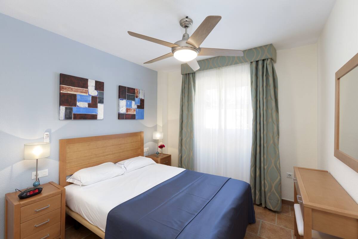 Una habitación de hotel con cama, escritorio, ventilador de techo y obras de arte en la pared.