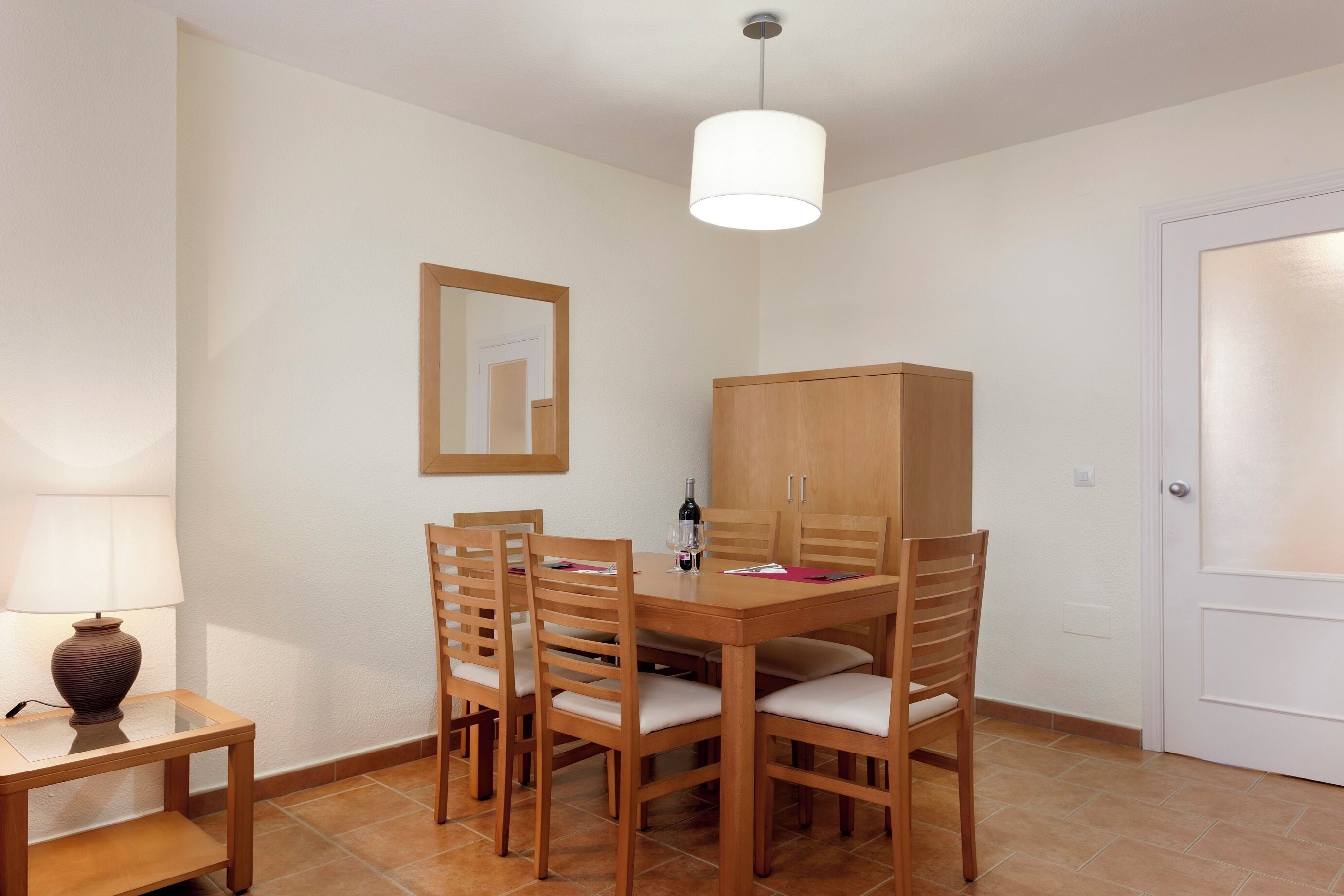 Een eetgedeelte met een houten tafel, vier stoelen, een kast, een lamp en een deur.