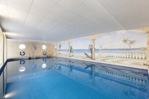 A large indoor swimming pool with clear water, surrounded by a decorative mural of a beach scene, including palm trees and a fence.