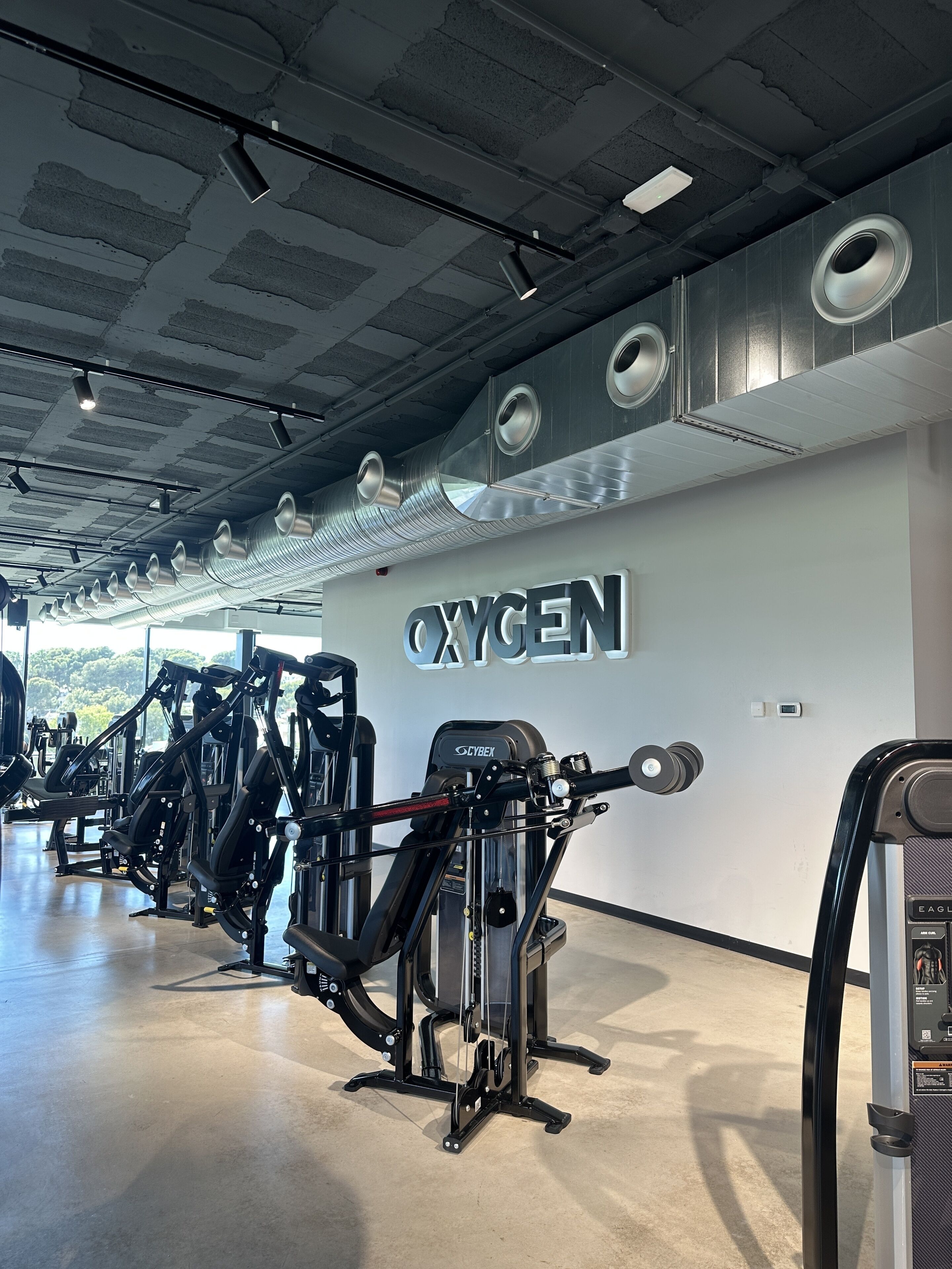 Un gimnasio con diversas máquinas de ejercicio, un techo con conductos de ventilación y una pared con la palabra 'OXYGEN'.