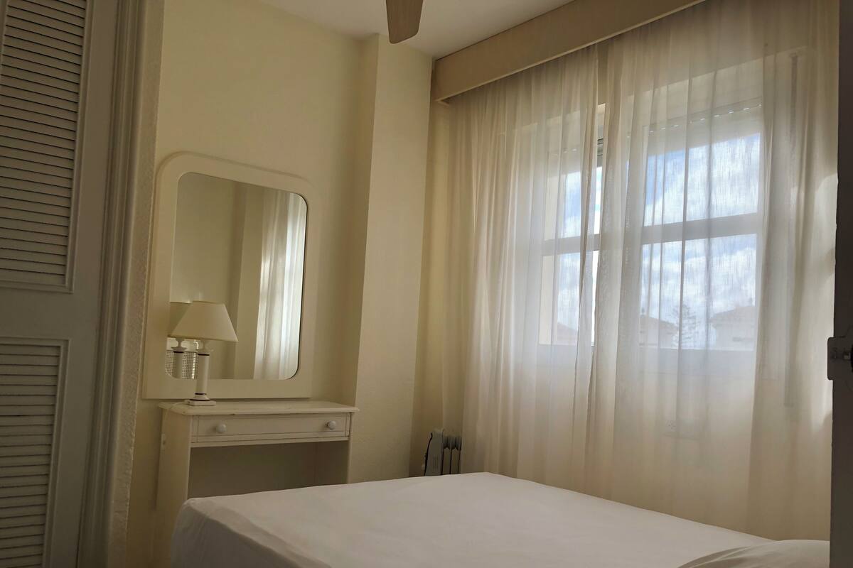 Una habitación de hotel con cama, mesita de noche con lámpara, espejo y cortinas traslúcidas sobre una ventana.