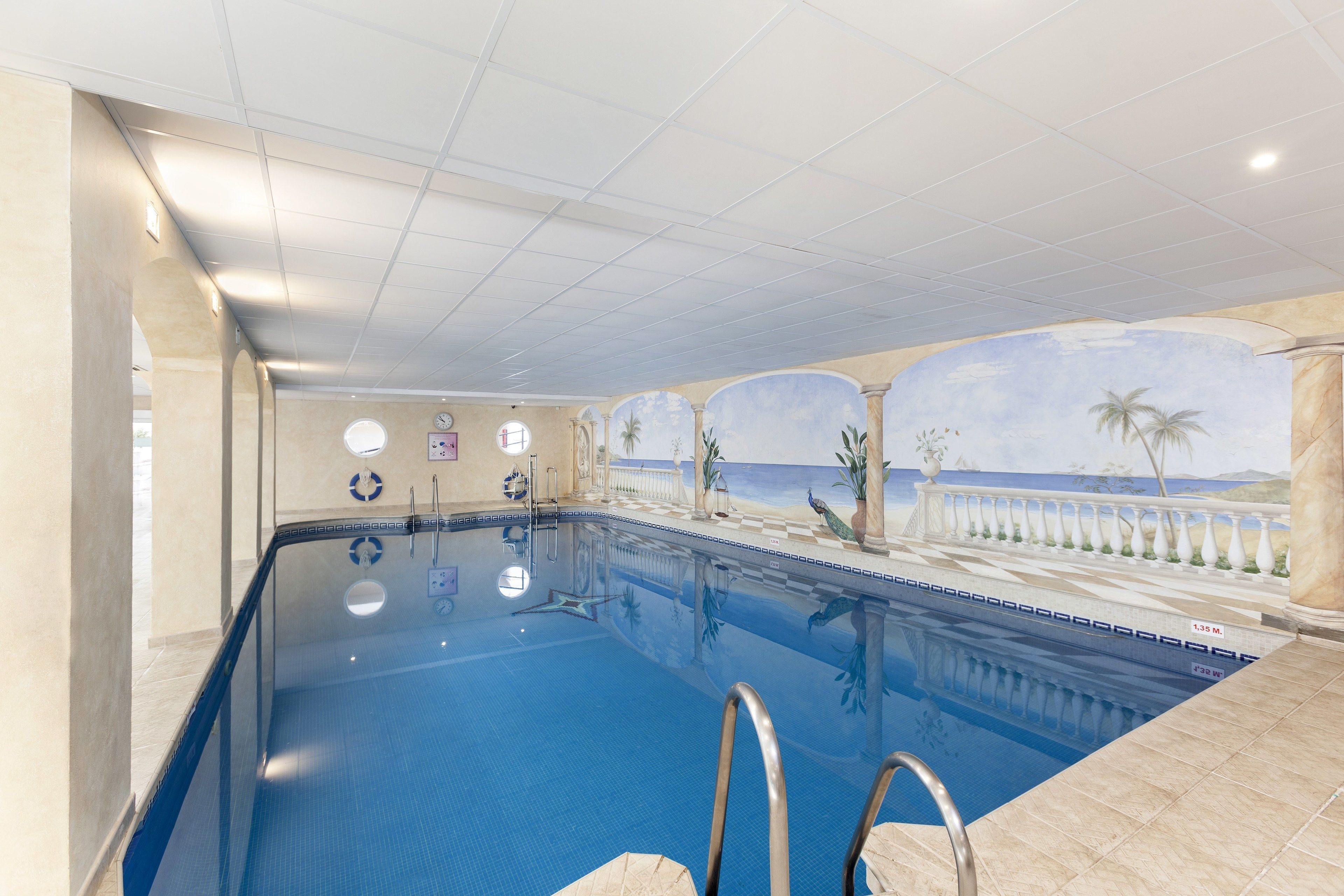 An indoor swimming pool with clear water, a tiled deck, and a mural of a beach scene.