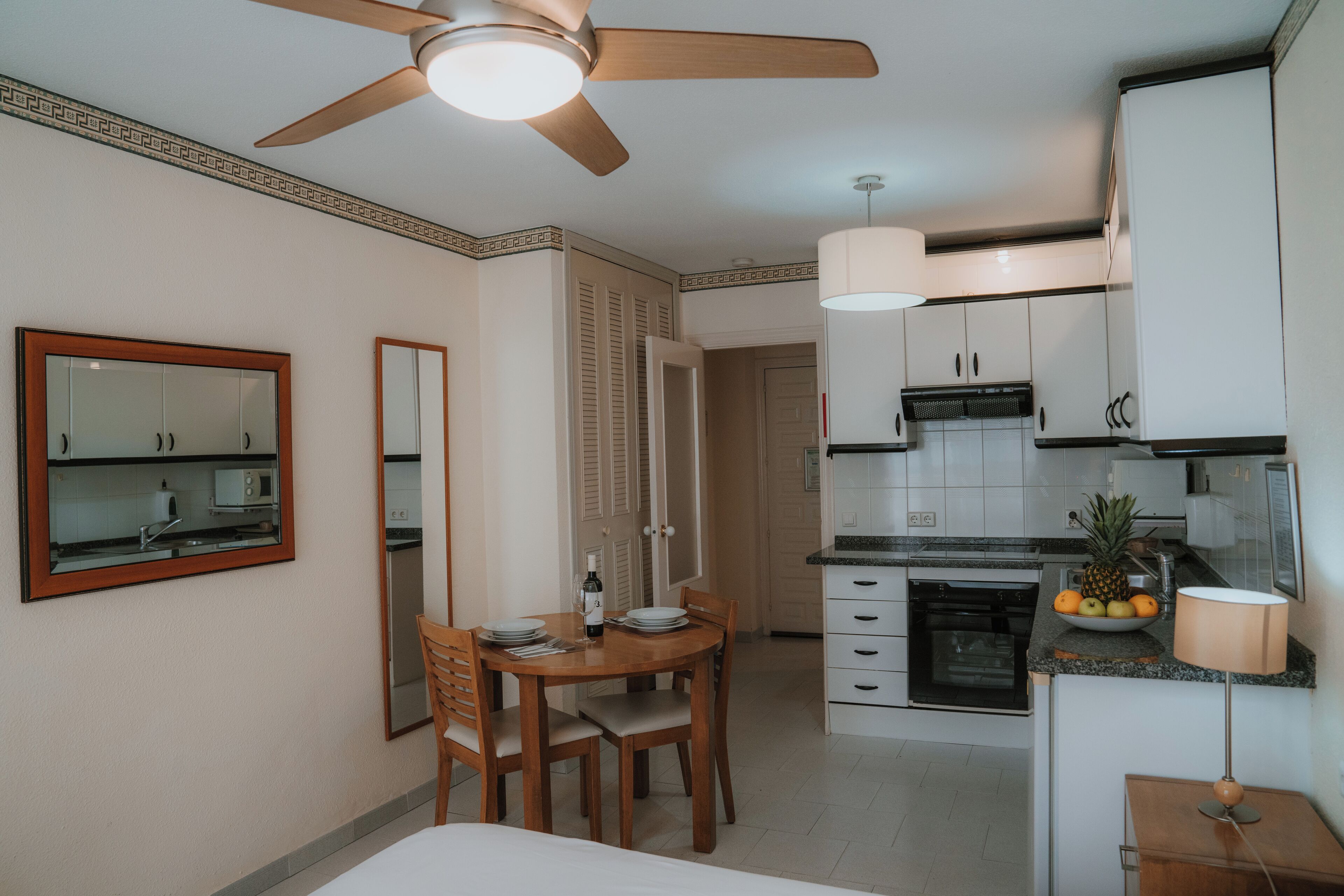 A compact kitchen with white cabinets, a dining area with a wooden table and chairs, a ceiling fan, and a microwave.