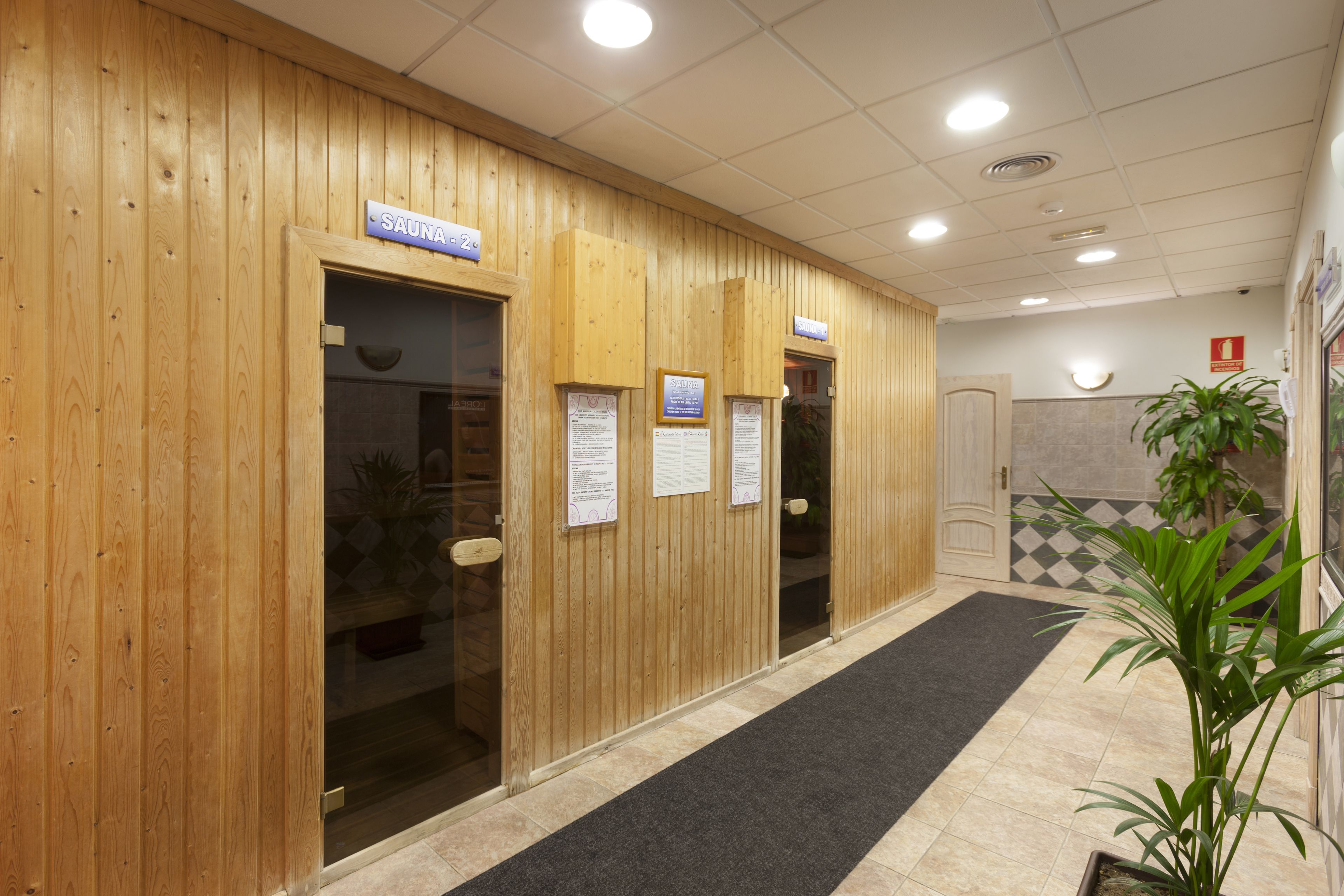Un espace d’accueil en bois, avec un panneau indiquant l’emplacement du sauna, un sol recouvert d’un tapis noir et une plante en pot.