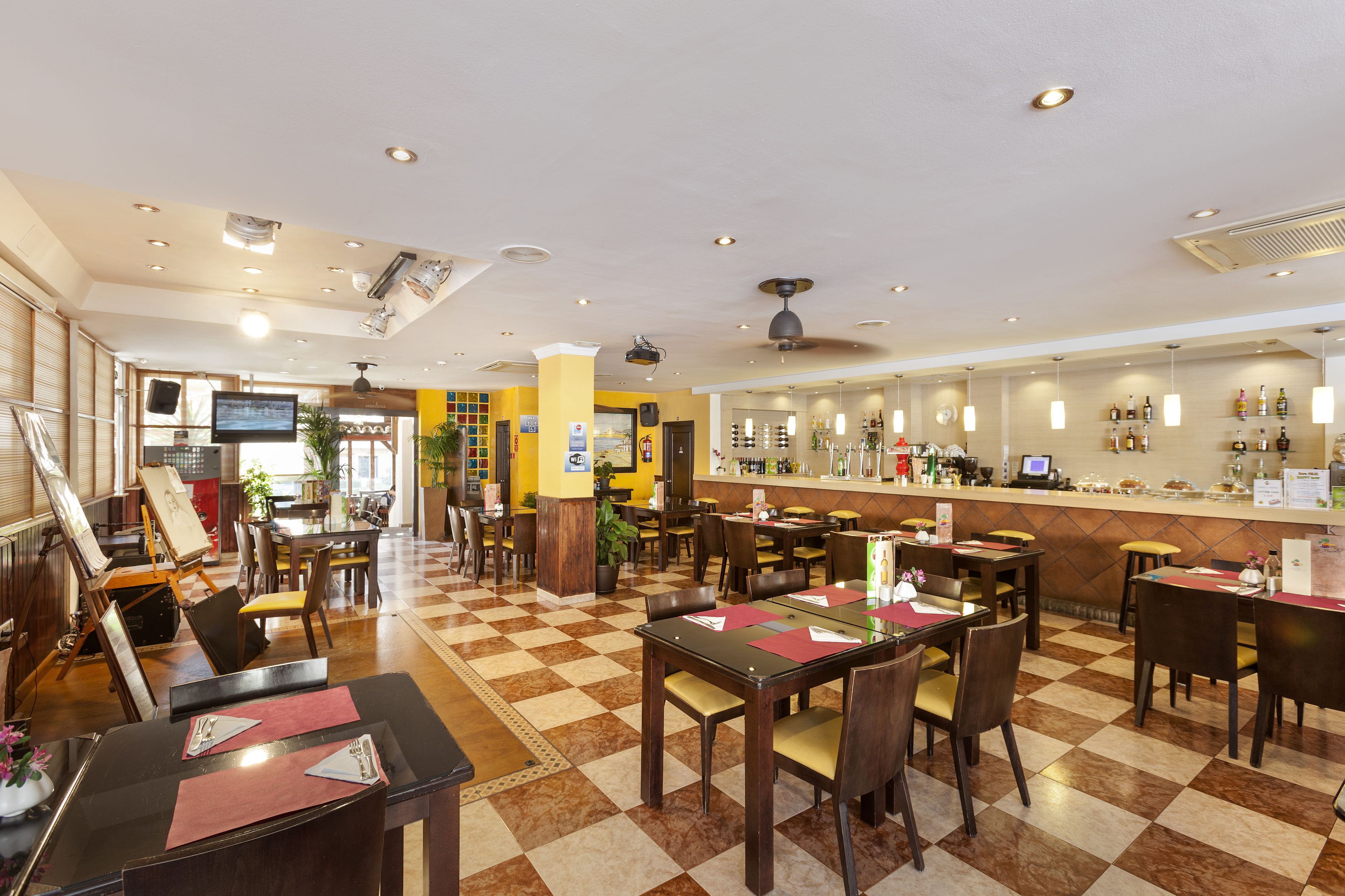 A hotel lobby with a checkered floor, multiple dining tables, chairs, a bar area, and a television screen.