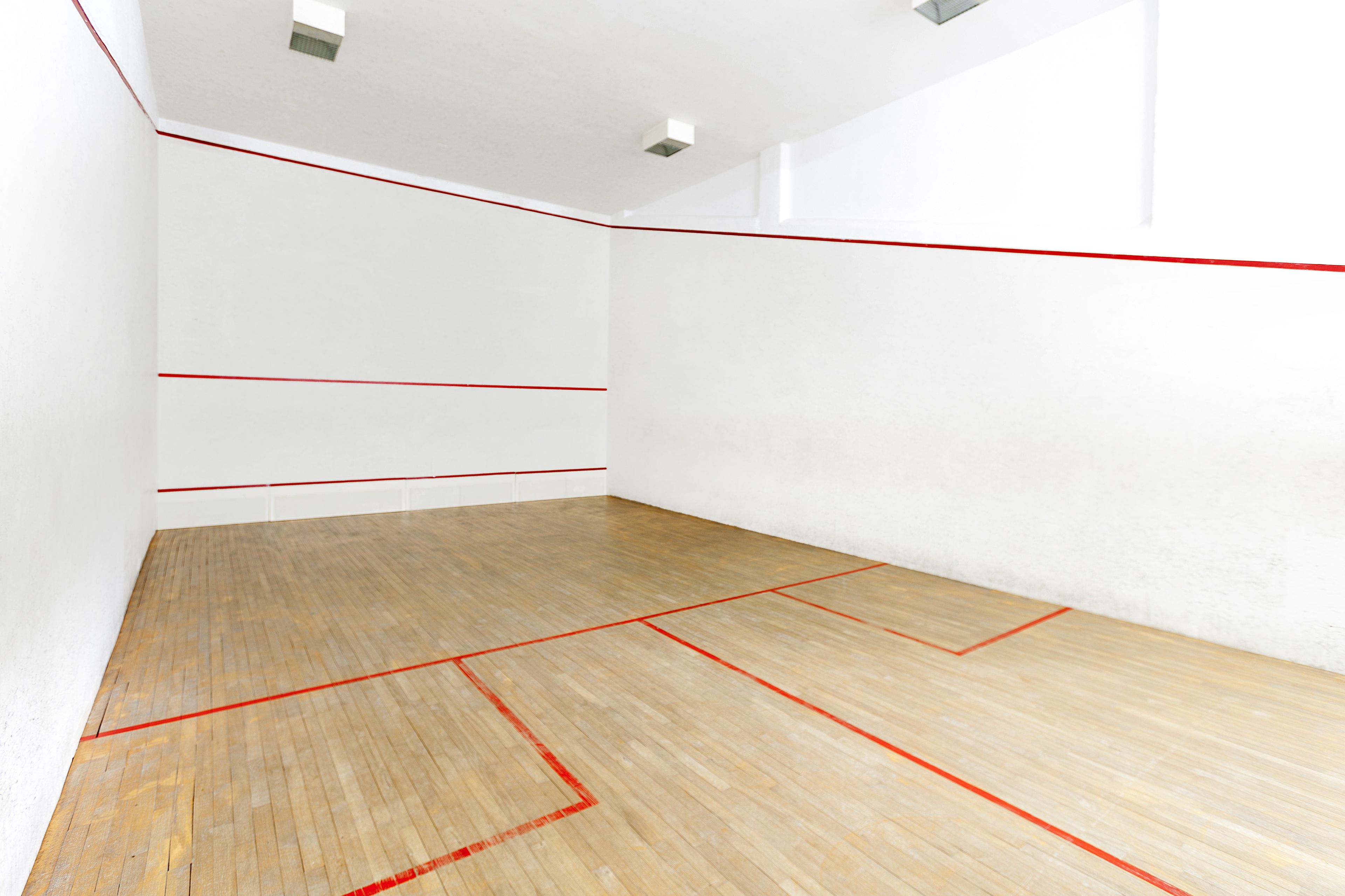 A squash court with white walls and a wooden floor marked with red lines.