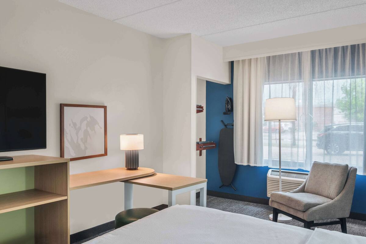 A hotel room with a bed, a desk with a lamp, a chair, a TV, and a window with curtains.
