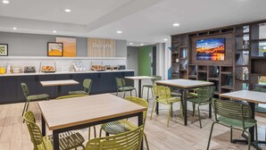 A modern cafe with wooden tables and green chairs, a counter with food items, and a TV on the wall. - Spark by Hilton St. Augustine Historic District (St. Augustine)