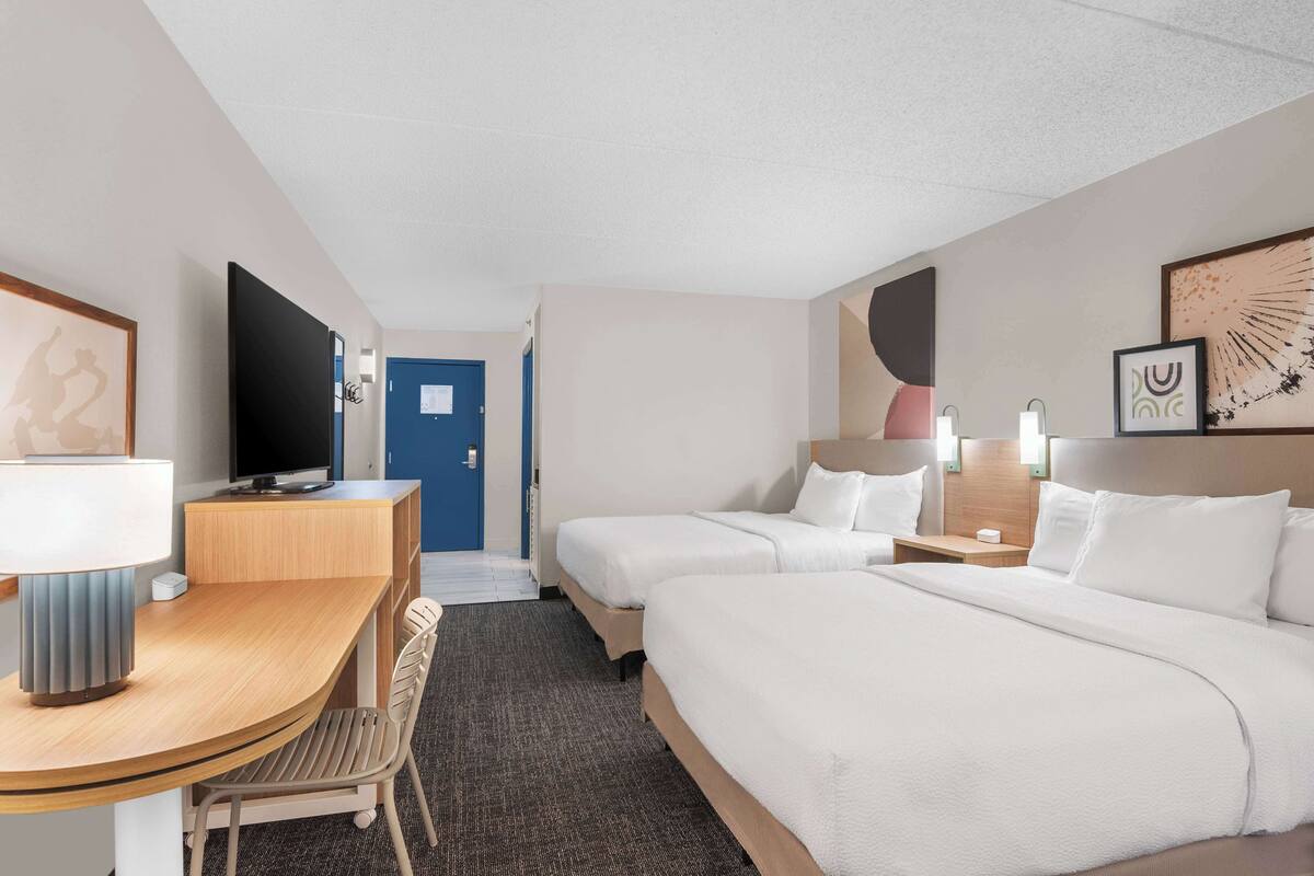 A hotel room with two beds, a desk with a chair, a TV, and a door leading to another room.