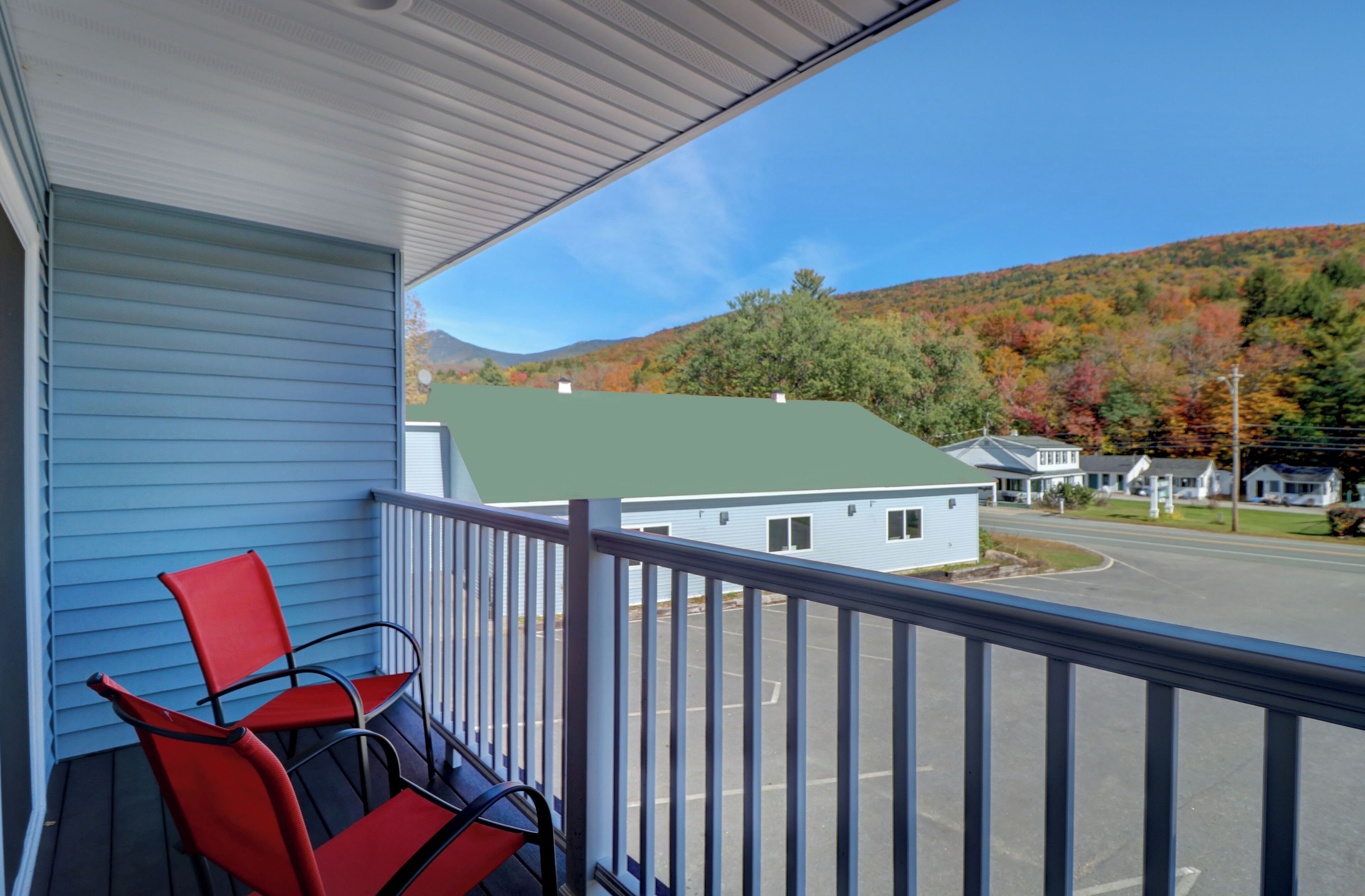 A balcony with two red chairs, a view of a street, and houses with a green roof in the background.