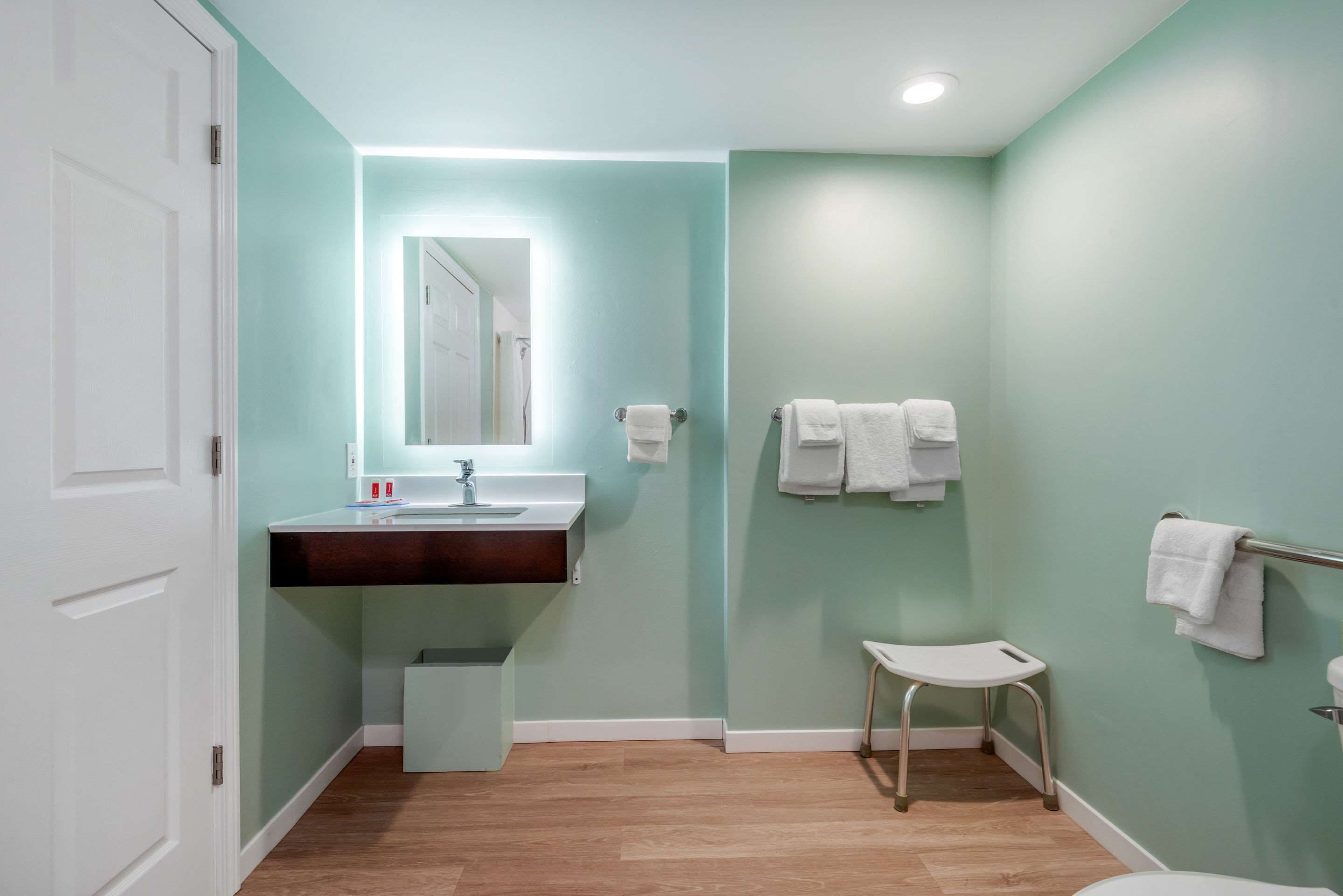A bathroom with a white door, a sink with a mirror, a small stool, and two towel racks.