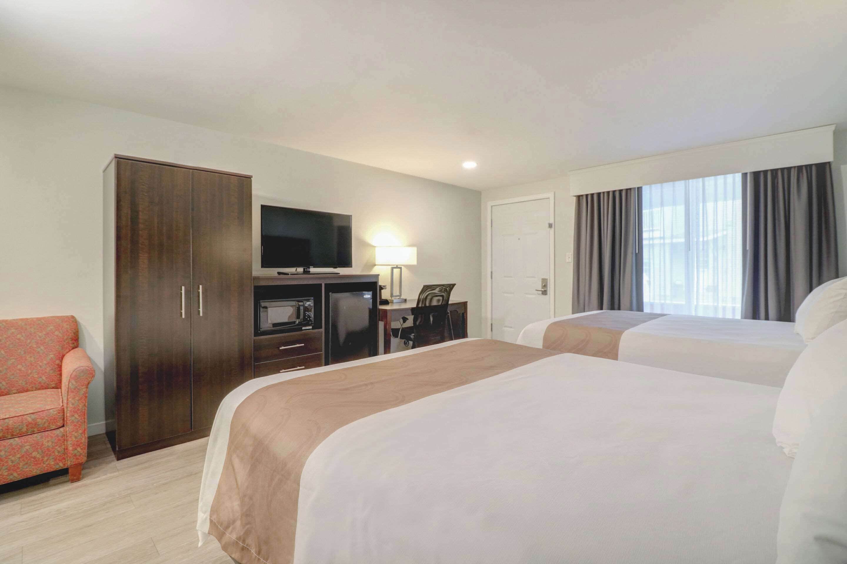Habitación de hotel con dos camas, televisión, escritorio con lámpara, armario y ventana con cortinas.