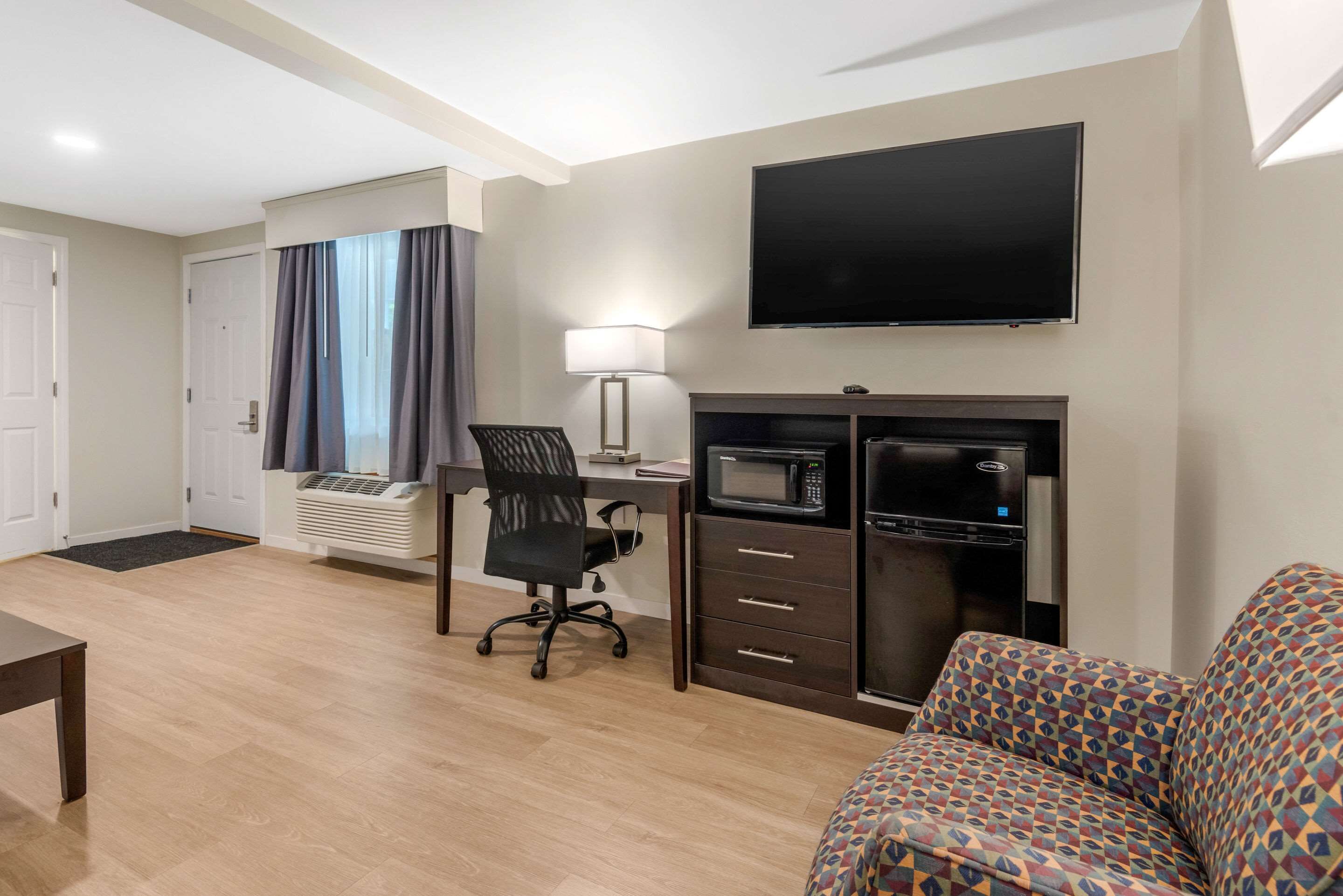 A hotel room with a flat-screen TV, a desk with a lamp, a microwave, a sofa, and a chair.