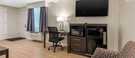A hotel room with a flat-screen TV, a desk with a lamp, a microwave, a sofa, and a chair.