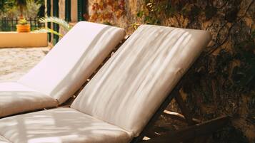 A sun lounger with a cushion, positioned against a stone wall with greenery, overlooking a beach and palm trees.