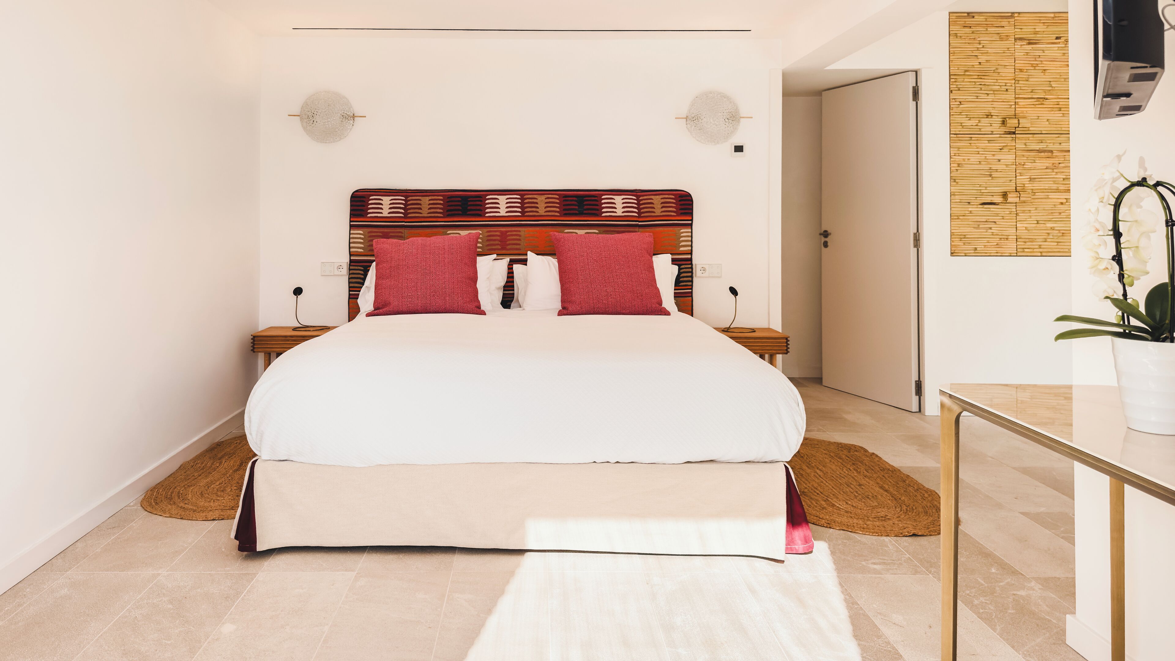 Una habitación de hotel moderna con una cama grande, cabecera de madera, dos mesitas de noche, una lámpara de noche, una pequeña mesa con una planta en maceta, y una puerta que da a otra habitación.