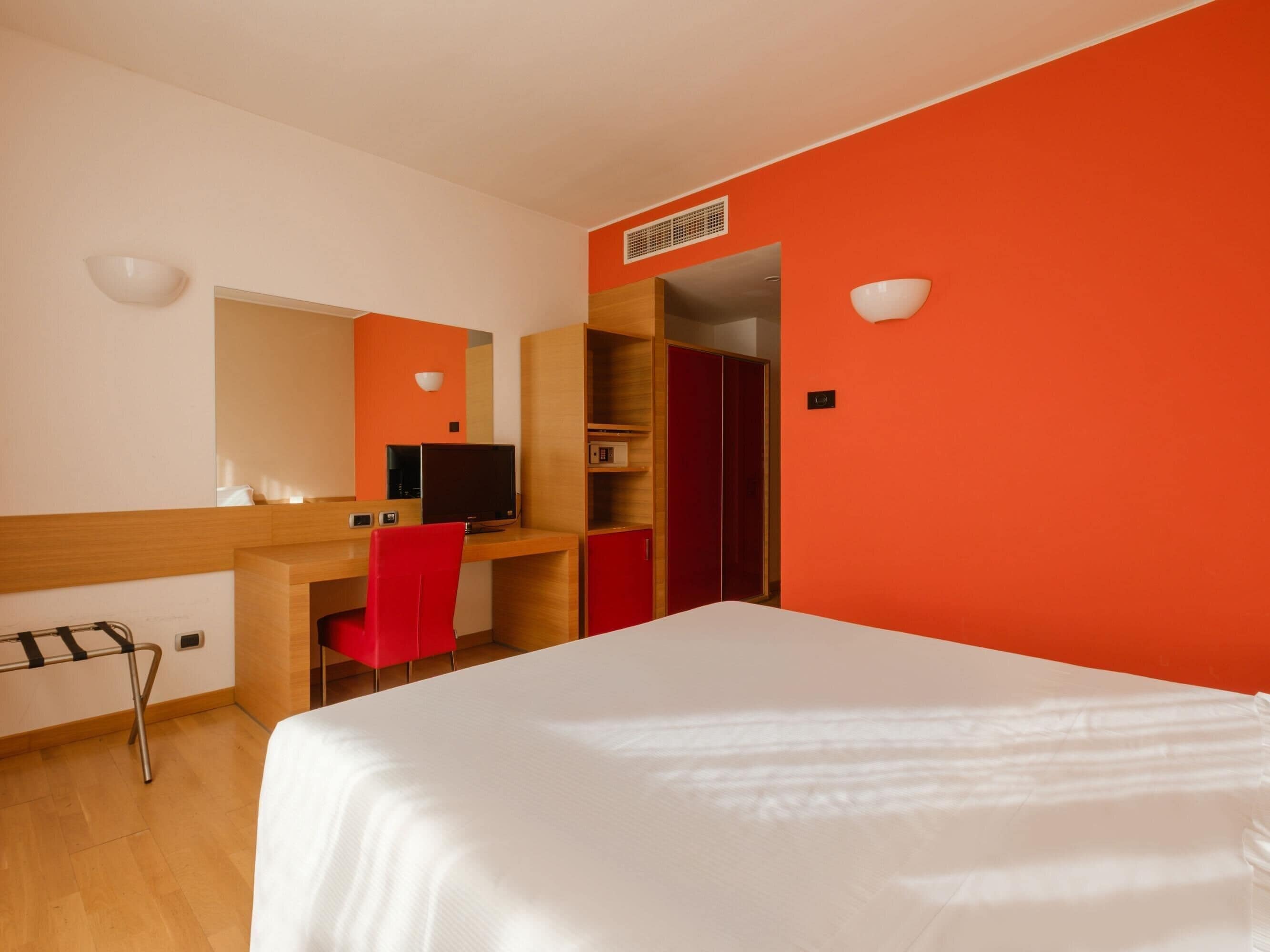 A hotel room with a bed, a desk with a computer, a red chair, and a wardrobe.