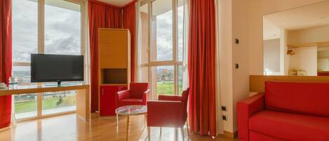Una sala de estar moderna con un sofá rojo, un sillón rojo, una mesita redonda y un televisor sobre un mueble de madera. Amplias ventanas con cortinas rojas ofrecen vistas al exterior.