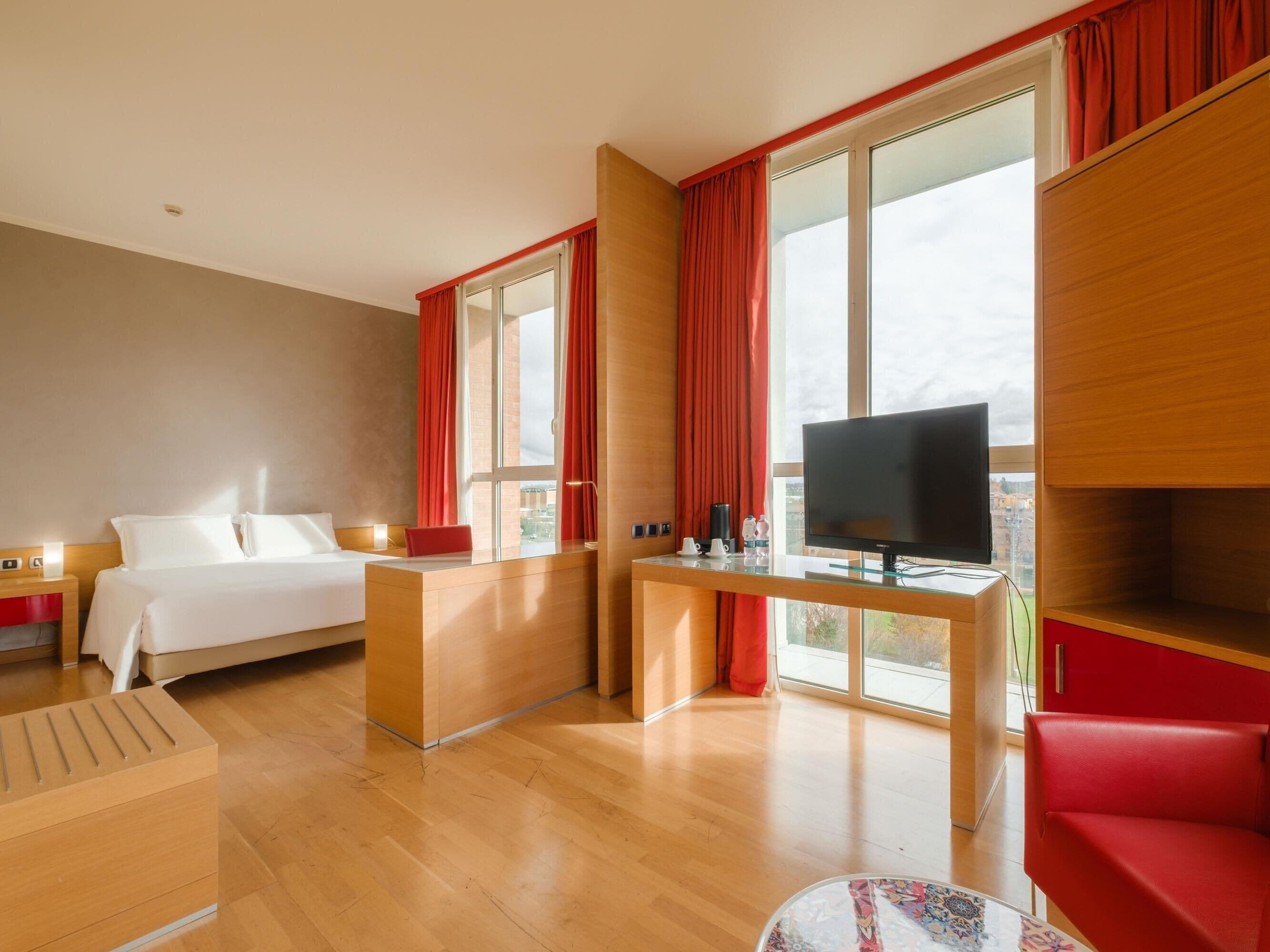 Una habitación de hotel moderna con una cama, un escritorio con televisor, una silla roja y ventanas amplias con cortinas.