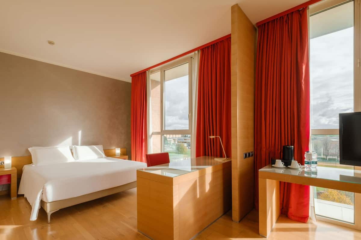 Una habitación de hotel con cama, escritorio, televisor y ventana con cortinas rojas.