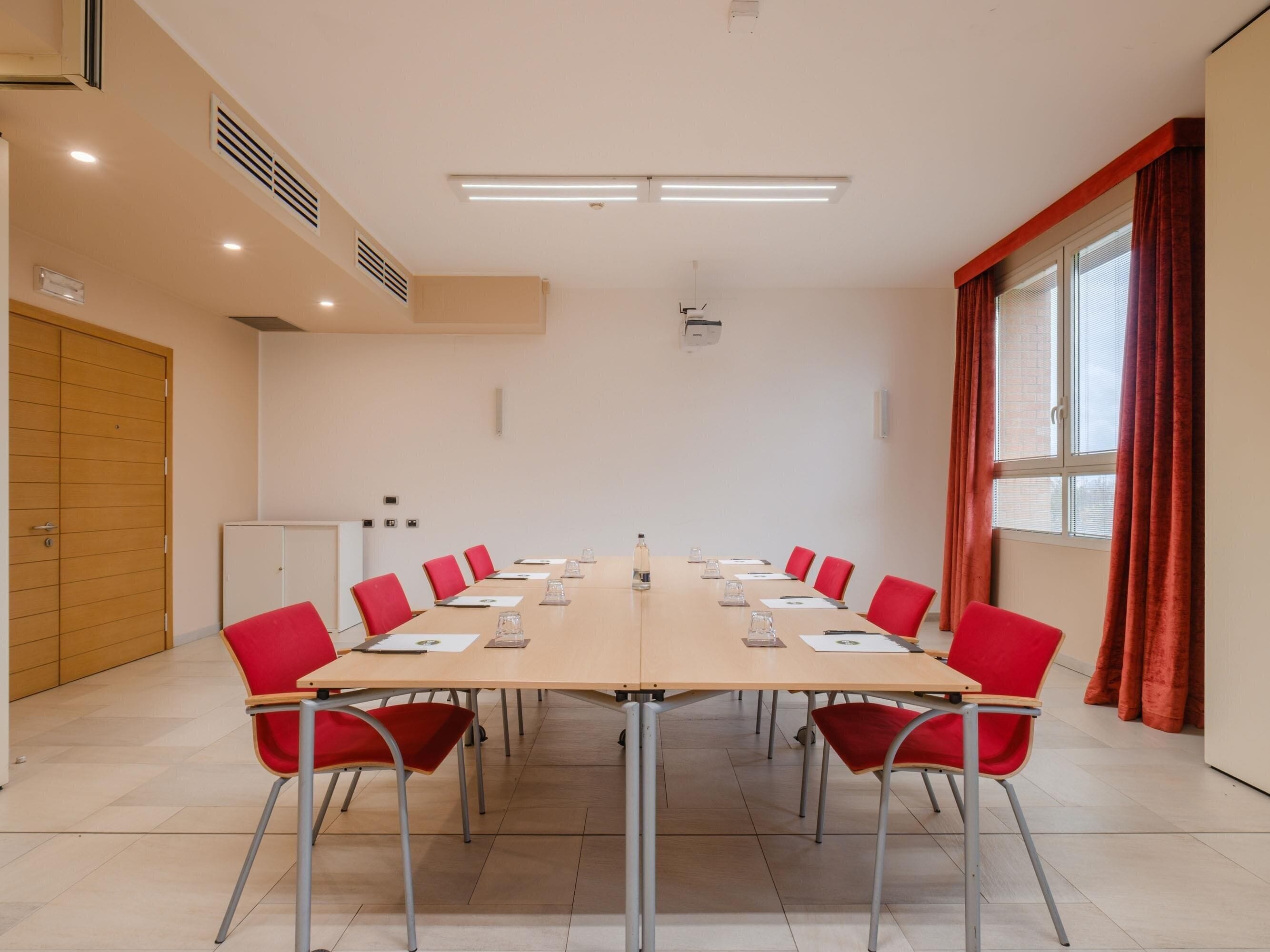 A conference room with a long table, red chairs, a whiteboard, and a window with curtains.