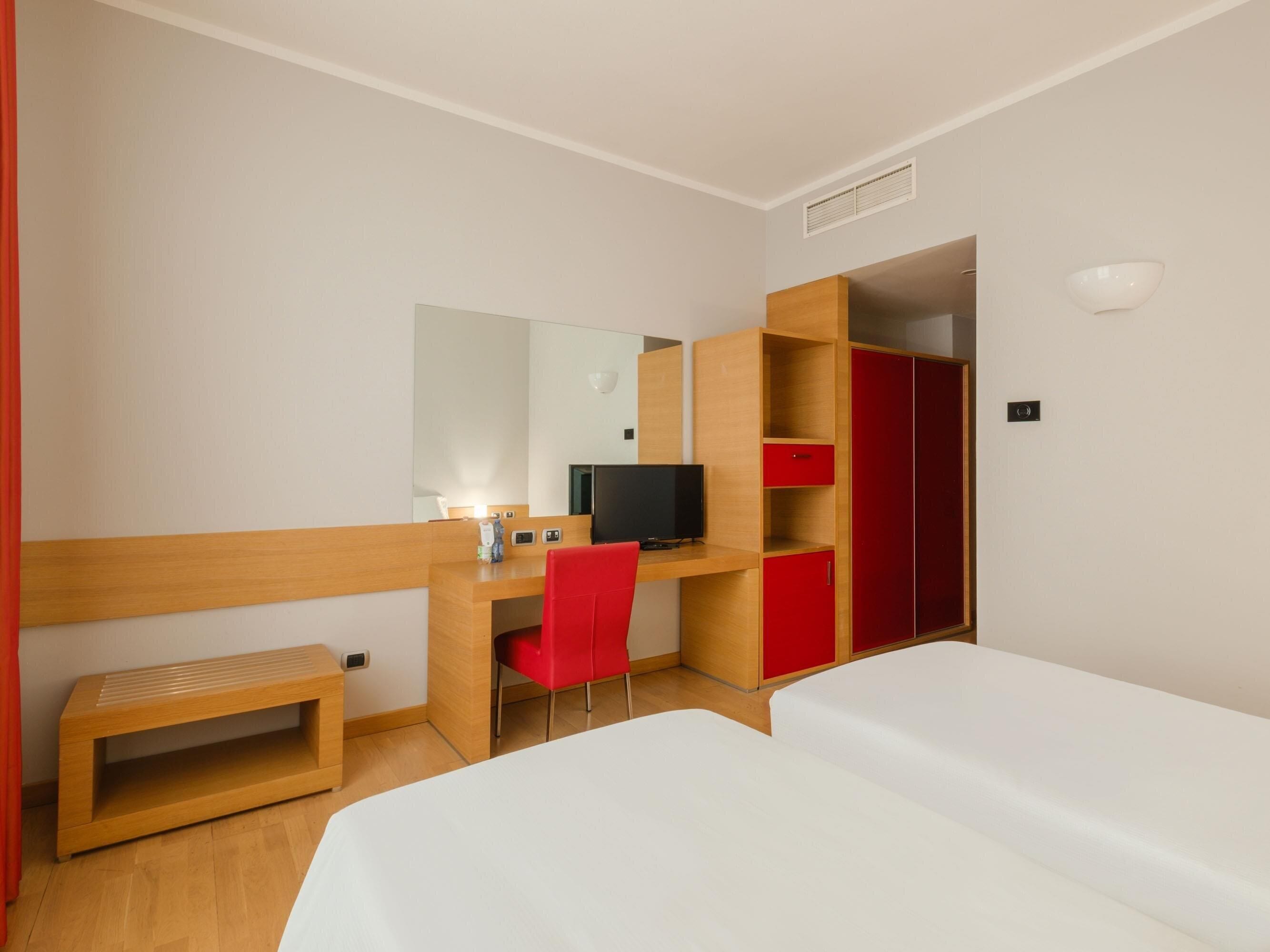 Ein Hotelzimmer mit Schreibtisch aus Holz, einem roten Sessel, einem Fernseher, einem Kleiderschrank und zwei Betten.