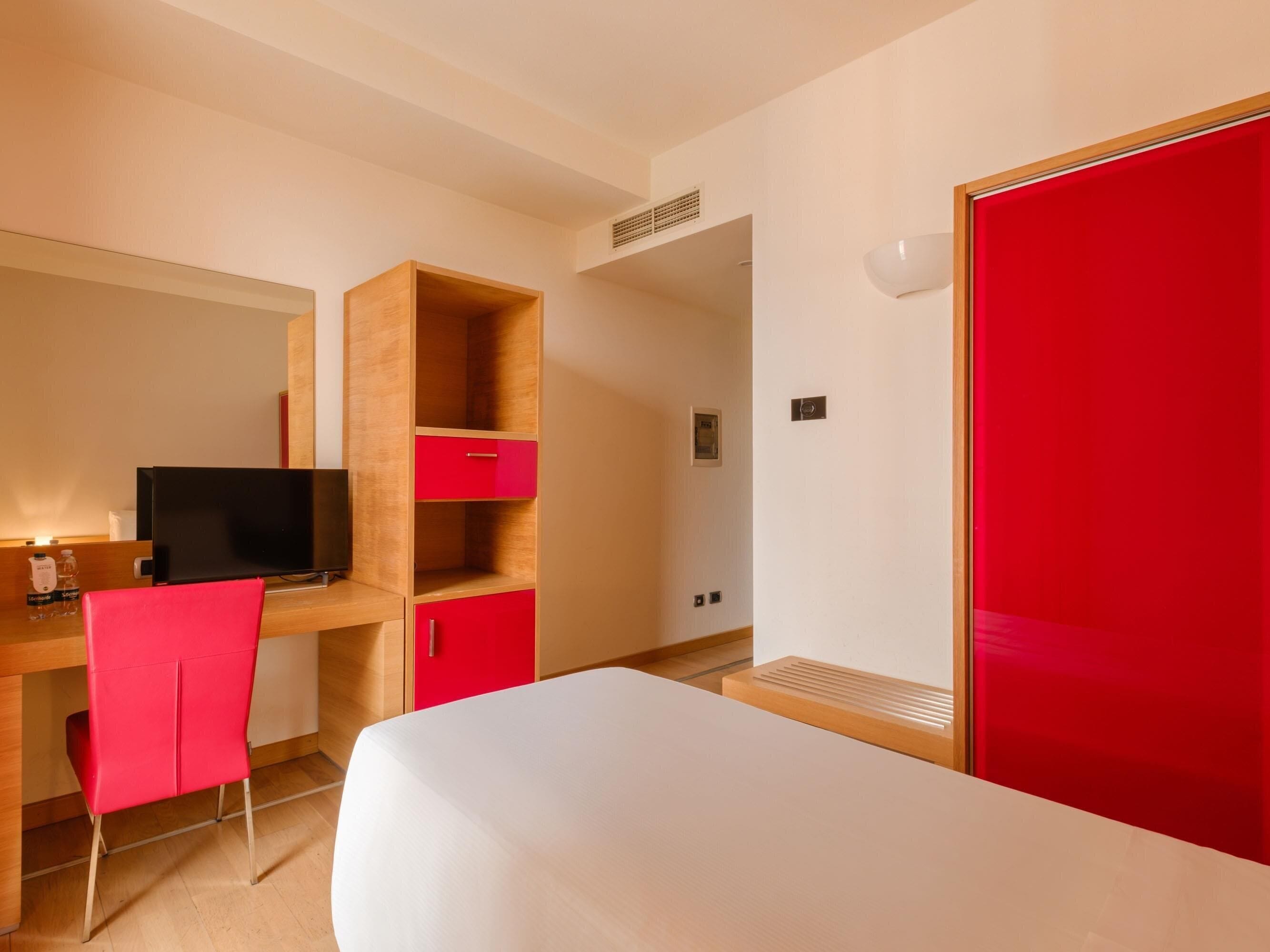 Quarto de hotel com cama, escrivaninha com computador, uma cadeira vermelha e guarda-roupa vermelho.