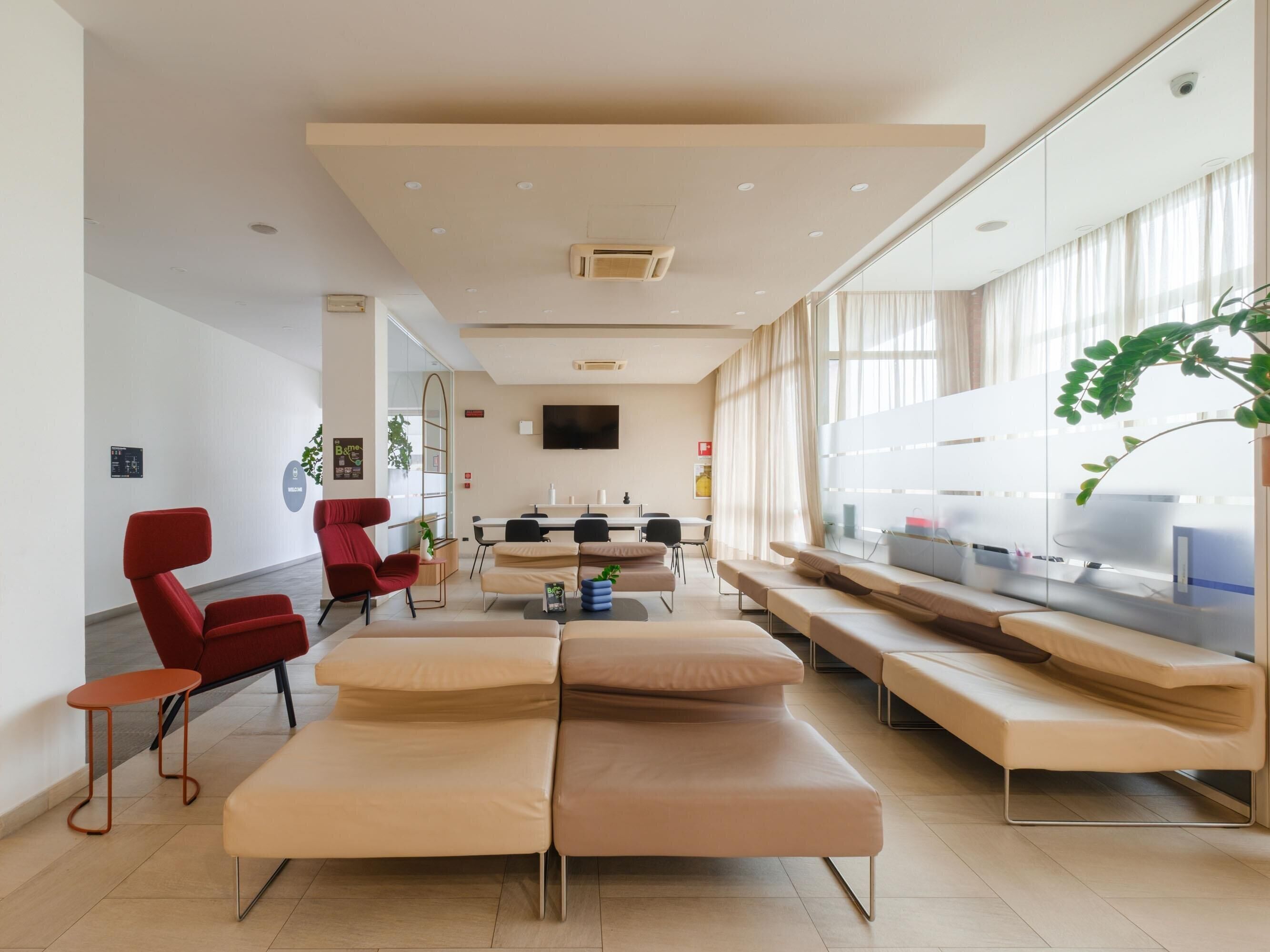 Un vestíbul d'hotel modern amb una recepció, zona de seients amb sofàs i cadires, una gran pantalla de televisió i una planta.