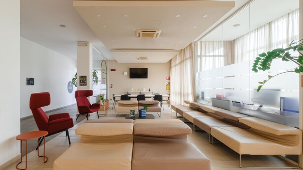 A modern hotel lobby with a reception desk, seating area with sofas and chairs, a large TV screen, and a plant. - B&B HOTEL Remilia (Reggio nell'Emilia)