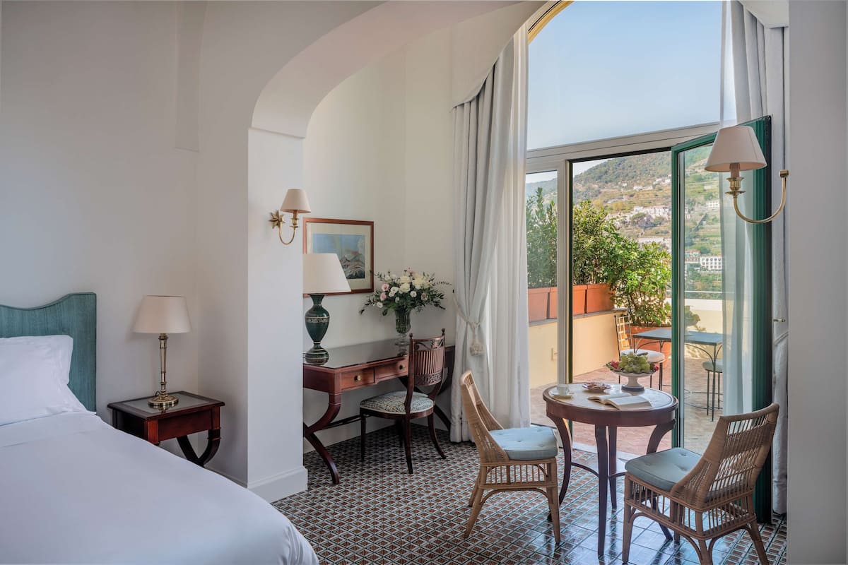 Une chambre d’hôtel comprenant un lit, un bureau avec une lampe, une chaise, une petite table avec un vase de fleurs et une vue sur l’extérieur grâce à une grande fenêtre.