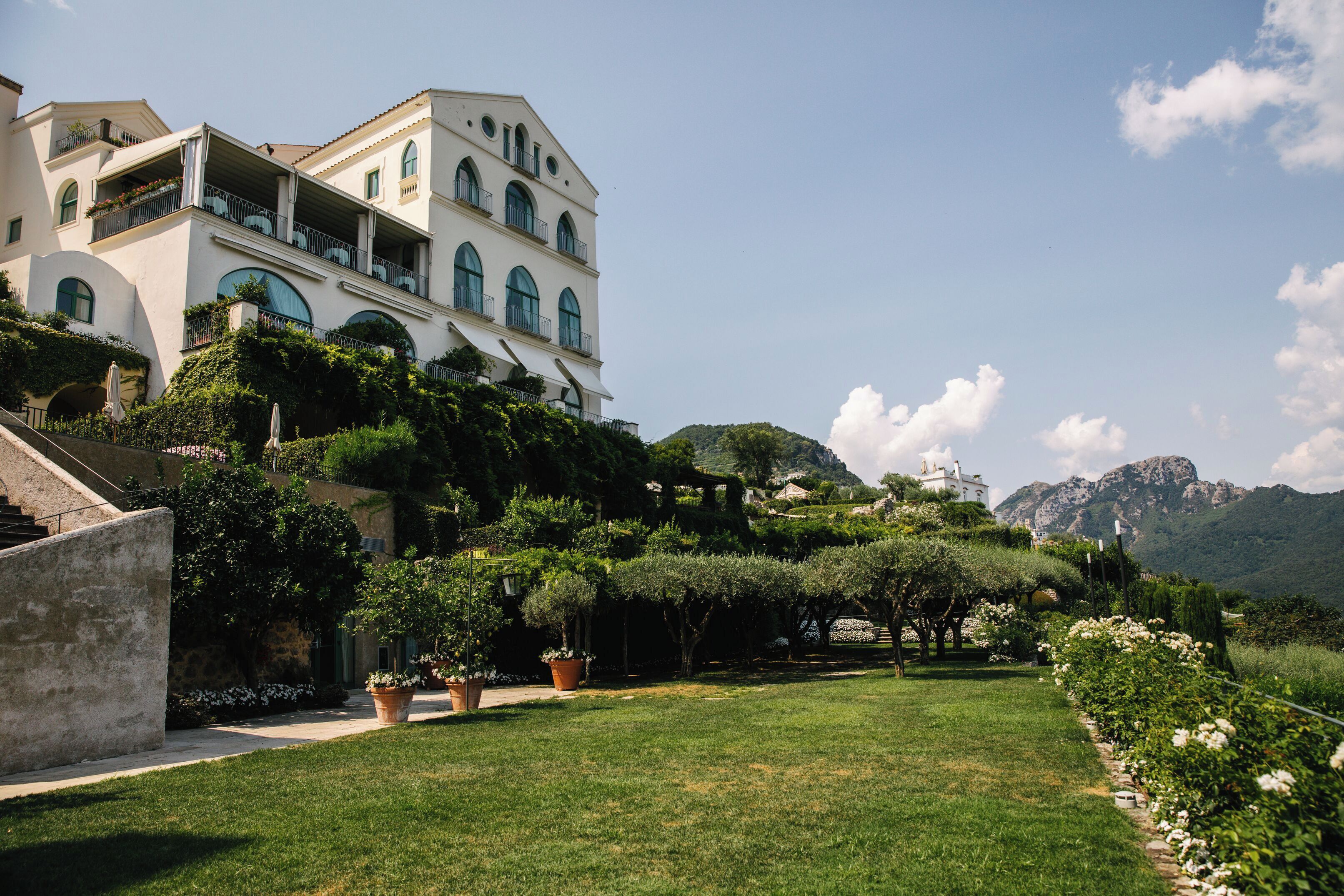 A large, multi-story building with balconies and arched windows, surrounded by lush greenery and a mountainous backdrop.
