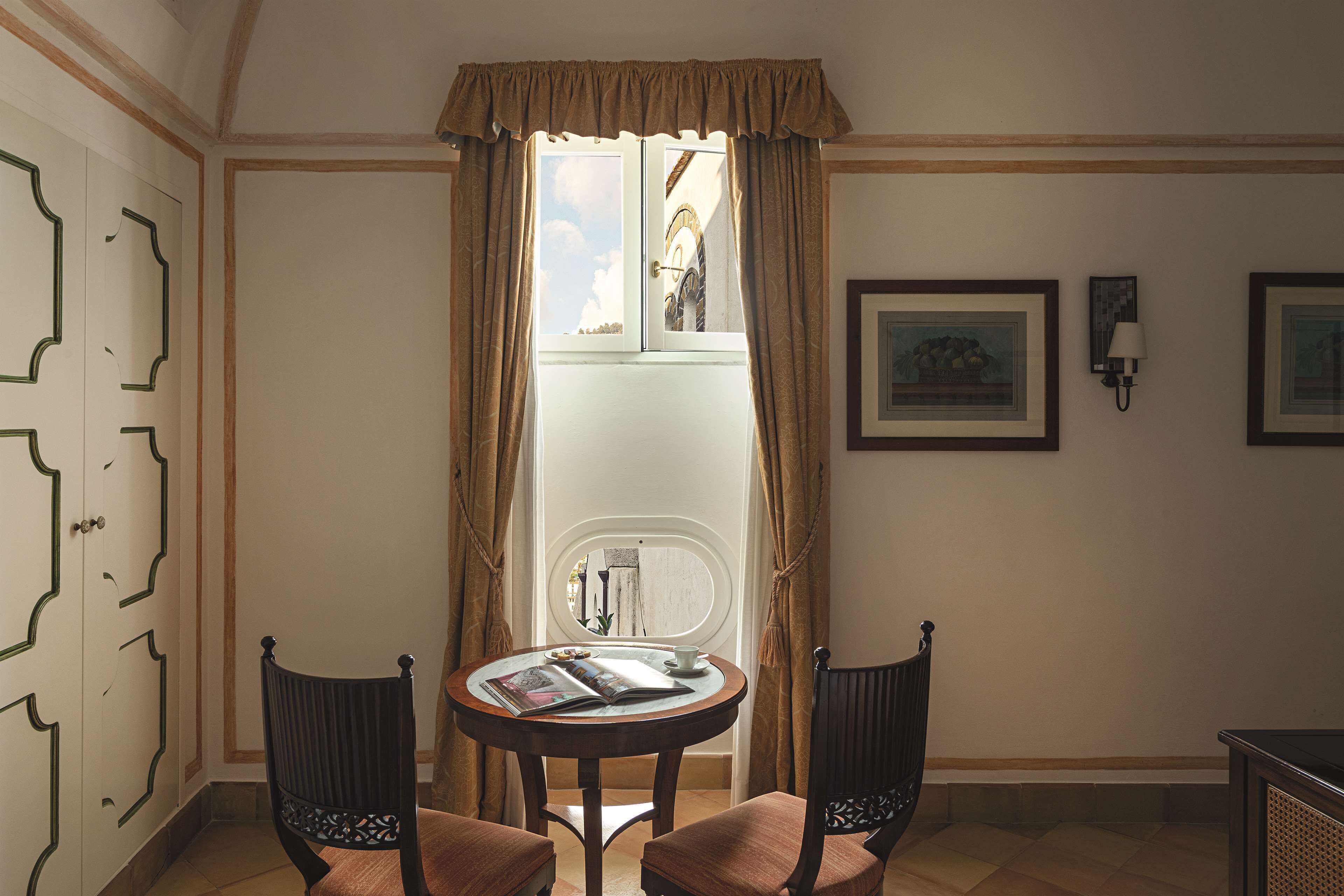 A room with a round table, two chairs, a window with curtains, a wall-mounted lamp, and framed pictures.