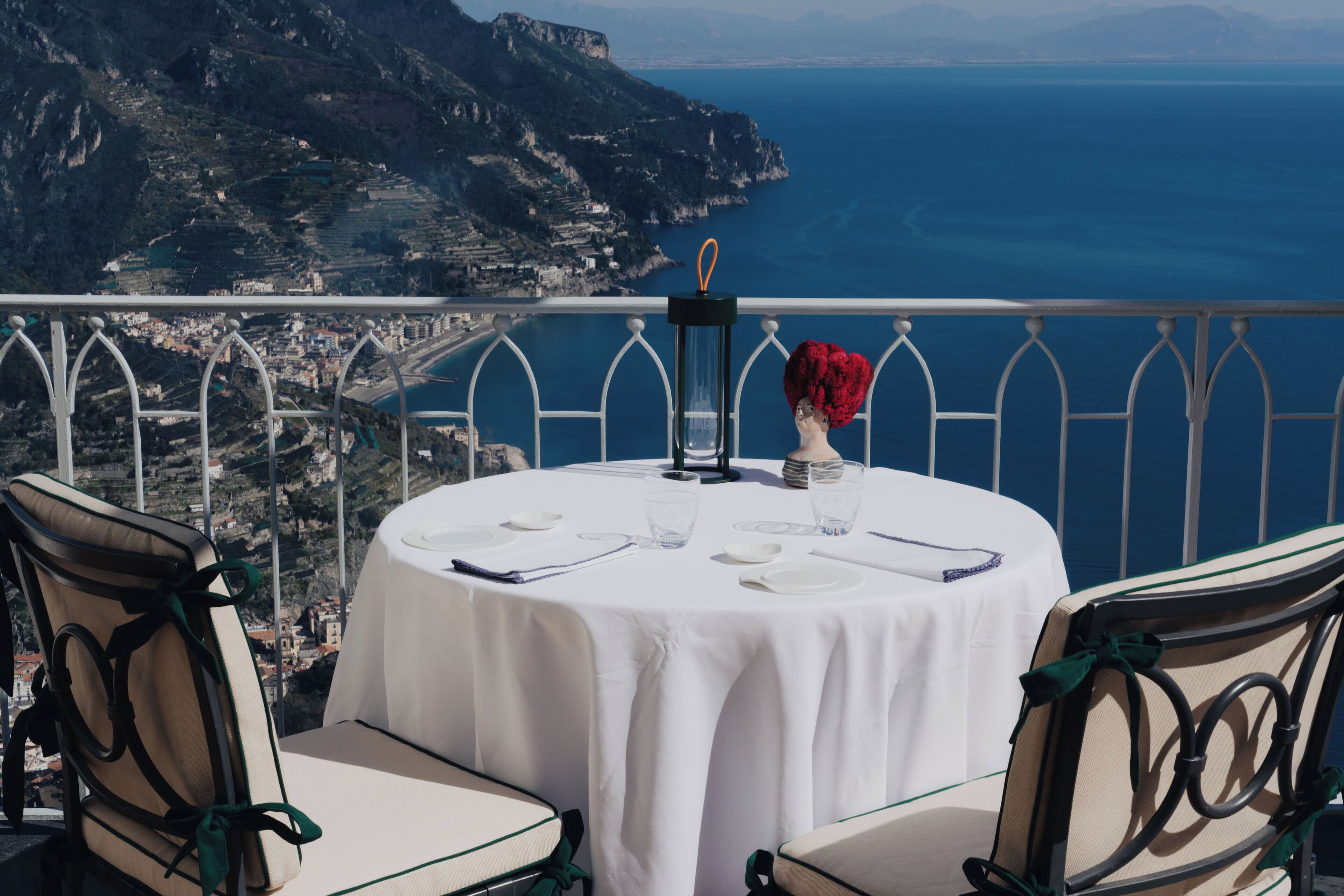 A round table with a white tablecloth, two chairs, a vase with red flowers, and a black candle holder. The table is set for two with plates, glasses, and cutlery.