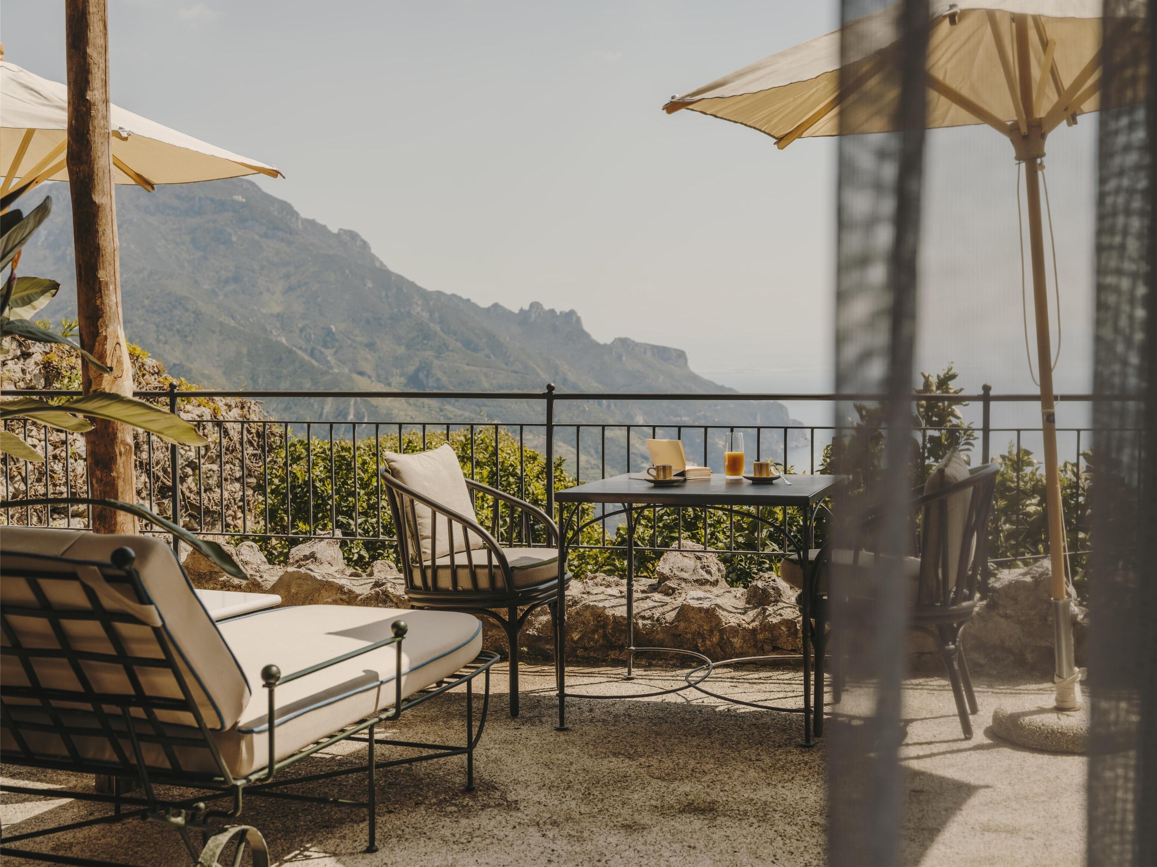 A terrace with a table, chairs, and umbrellas, overlooking a mountainous landscape.