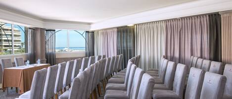 Un salón de conferencias con filas de sillas tapizadas en gris y una mesa con botellas de agua.